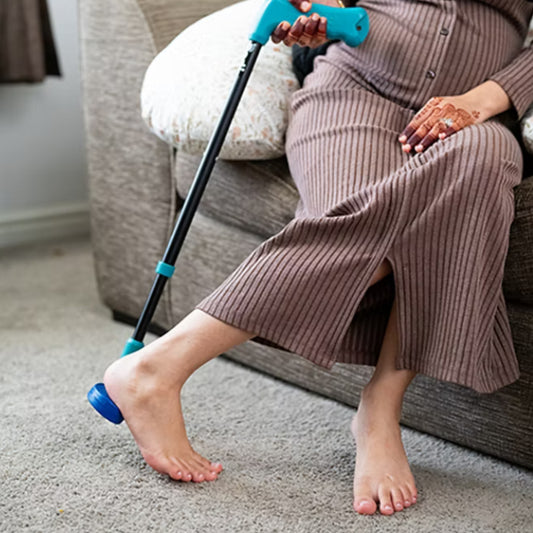 Person sitting on a sofa using the PediStick telescopic handle to reach and apply cream to their heel without bending.