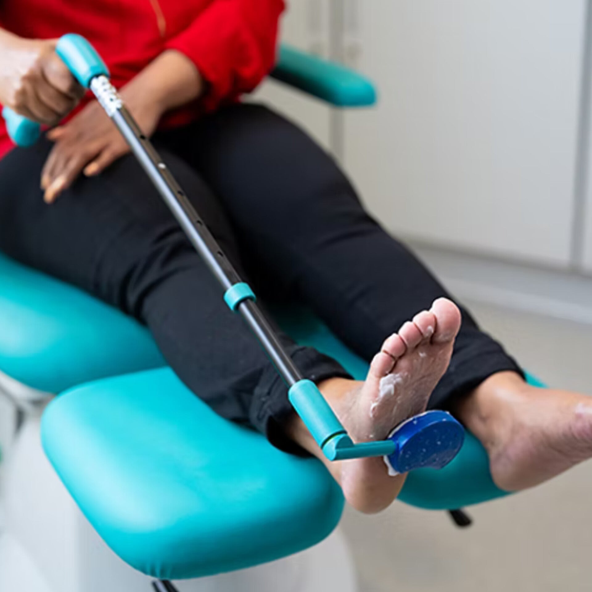 Person sitting in a medical chair using the PediStick long-handled applicator to apply foot cream to the sole of their foot.