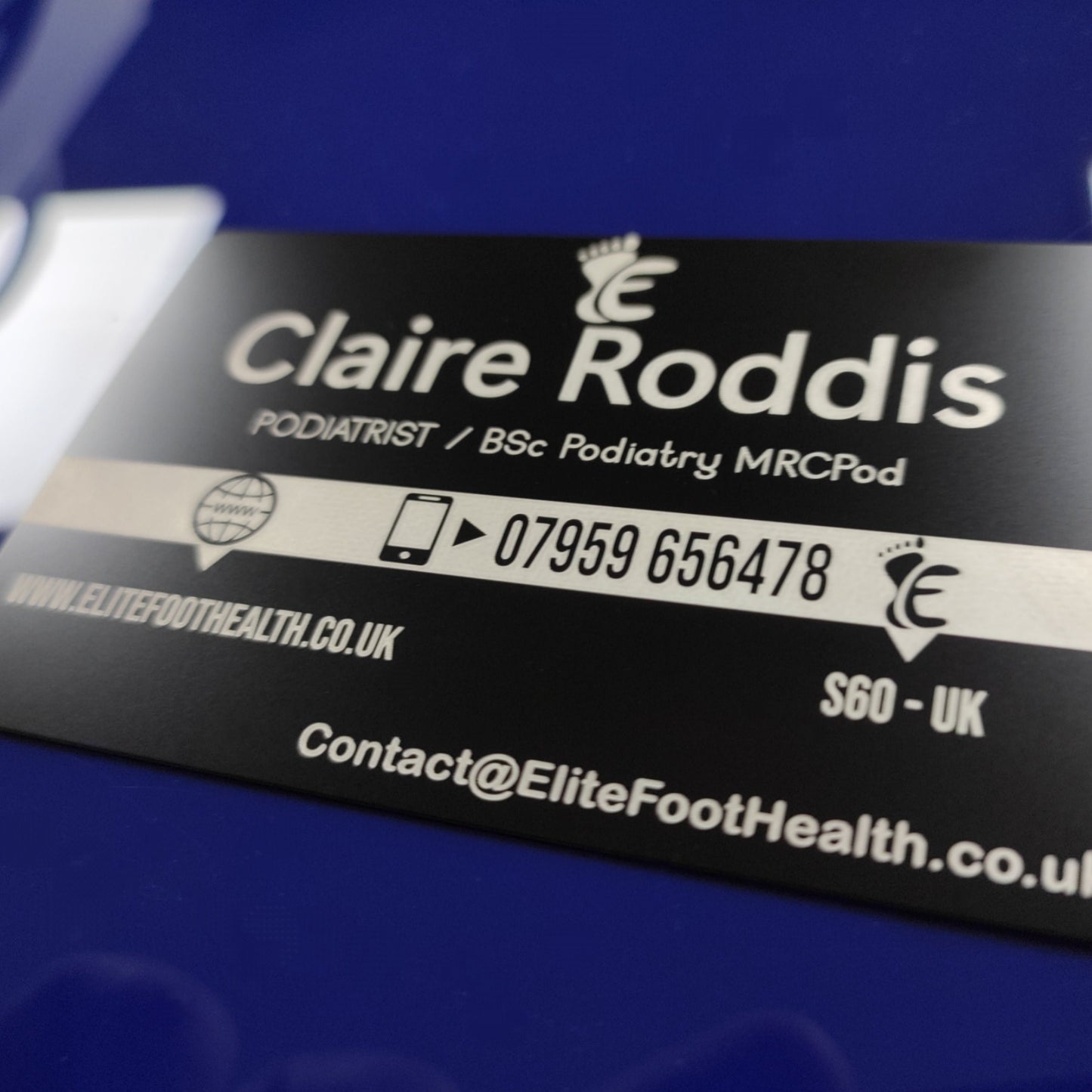 A black metal business card with white text, lying on a blue surface. The text includes a podiatrist's details and a contact number, with a design symbol and 'Elite Foot Health' branding.