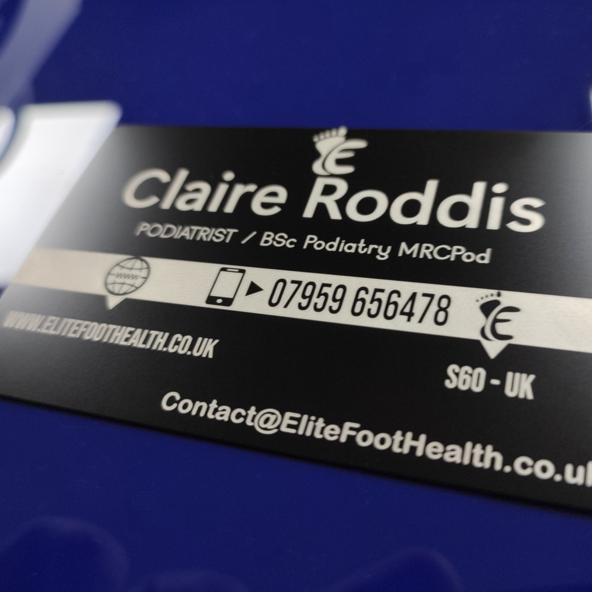 A black metal business card with white text, lying on a blue surface. The text includes a podiatrist's details and a contact number, with a design symbol and 'Elite Foot Health' branding.