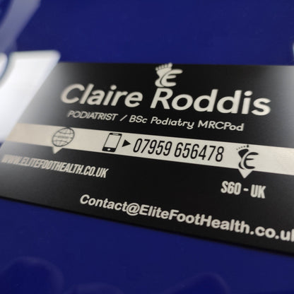 A black metal business card with white text, lying on a blue surface. The text includes a podiatrist's details and a contact number, with a design symbol and 'Elite Foot Health' branding.